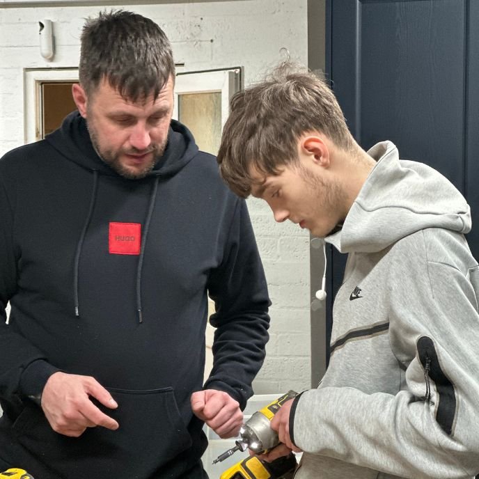 A mentor guides a young person using a drill in a workshop
