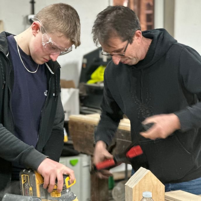 A young person sanding wood alongside a mentor
